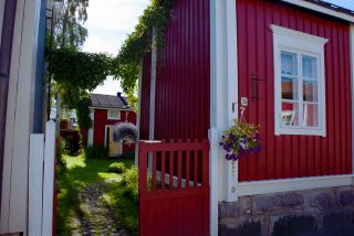 Kuvassa Kokkolan vanhakaupunki Neristanissa sijaitsevan punamultavärillä maalatun puutalon punainen portti joka johdattaa tulijansa vehreälle sisäpihalle. Kaunis kukkaamppeli roikkuu portin pielessä