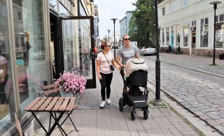 Perhe viettää kesäistä päiväänsä kävellen Kokkolan keskustassa mukulakivikadulla. Kivijalkaliikkeet ovat auki ja kaupan edessä on kukkia.